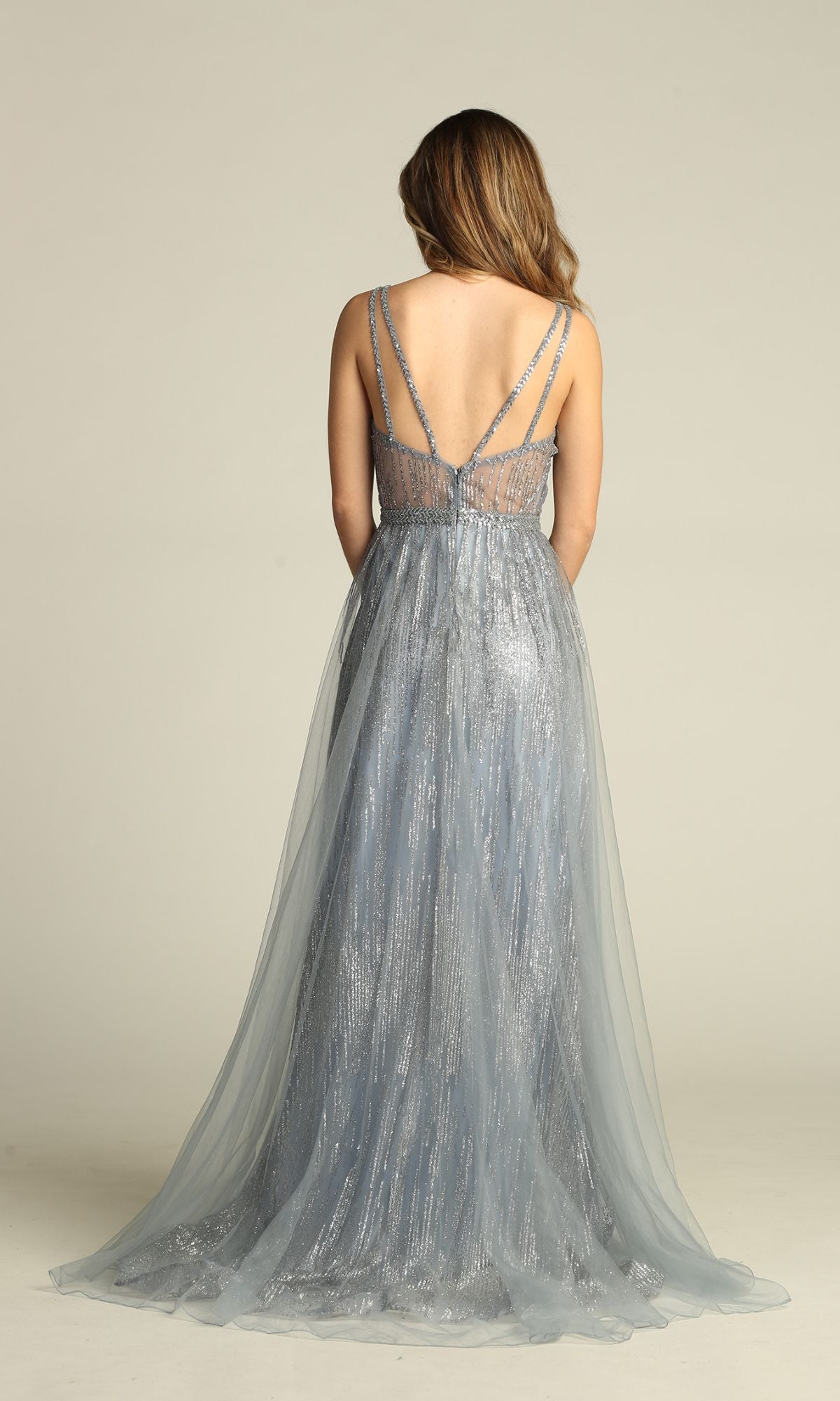 A woman stands with her back to the camera in the Sheer-Waist Long Sequin Prom Dress: Chicas YG5021, a sleeveless, floor-length gown with spaghetti straps and silver-blue shimmer that sparkles under the light against a plain backdrop.